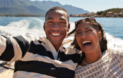 Ein lachendes junges paar sitzt auf einem Boot. Der Arm des Mannes ist um die Schulter der Frau gelegt.